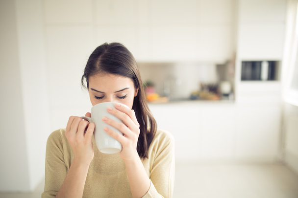 Young woman enjoying,holding cup of hot beverage,coffee or tea in morning sunlight.Enjoying her morning coffee in the kitchen.Savoring a cup of coffee breathing in the aroma - Фото, изображение