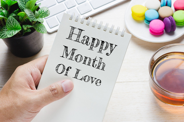 Man holding Happy month of love message on book and keyboard with a hot cup of tea, macaroon on the table. Can be attributed to your ad. - Photo, Image