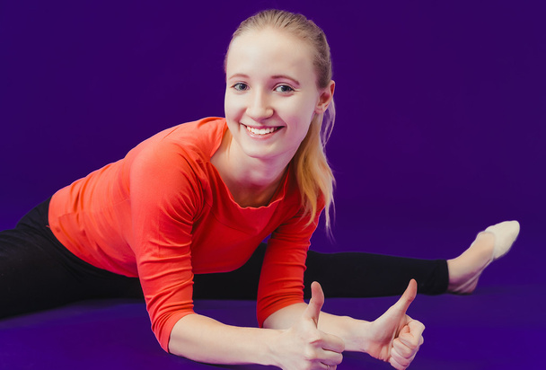 on fitness Smiling woman showing a thumbs up sign on a blue background sitting on the splits. Looking at camera - 写真・画像