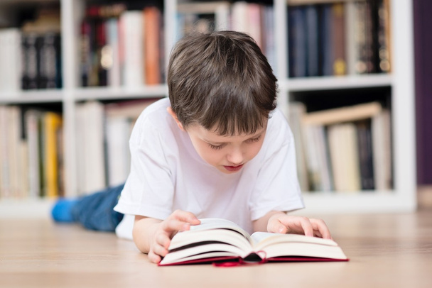 Αγόρι βρίσκεται στο στομάχι του και διαβάζοντας ένα βιβλίο στη βιβλιοθήκη - Φωτογραφία, εικόνα