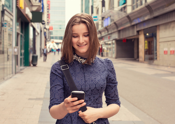 Portrait happy girl walking and texting on the smart phone in the street. Urban city background.     - Zdjęcie, obraz