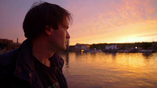 νεαρός άνδρας κοιτάζοντας το ηλιοβασίλεμα στο λιμάνι - Πλάνα, βίντεο