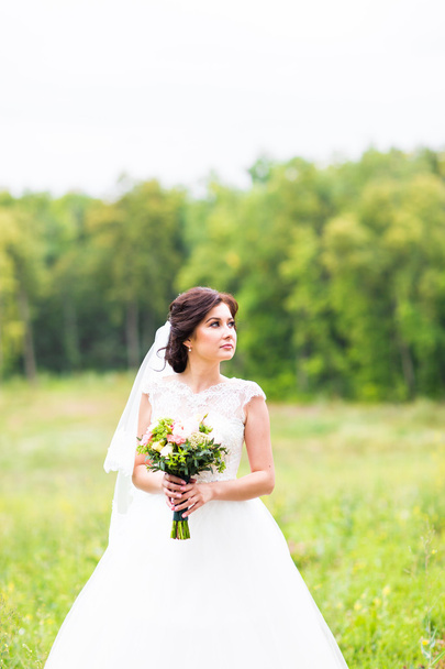 Beauty bride in bridal gown with bouquet and lace veil on the nature. Beautiful model girl in a white wedding dress. Female portrait in the park. Woman with hairstyle. Cute lady outdoors. - Foto, Imagen