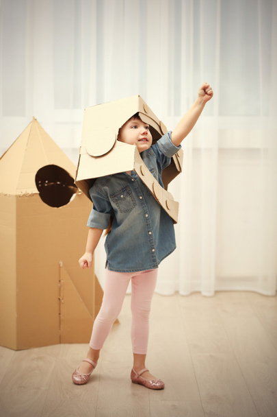 Little cute girl in astronaut helmet playing with cardboard space rocket in room - Photo, Image