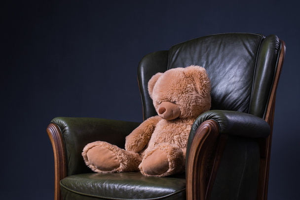 Teddy bear sits in a green leather armchair in front of the wall - Zdjęcie, obraz