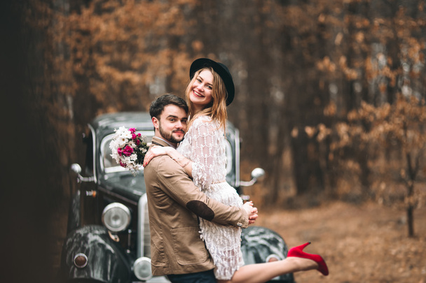 Gorgeous newlywed bride and groom posing in pine forest near retro car in their wedding day - Valokuva, kuva