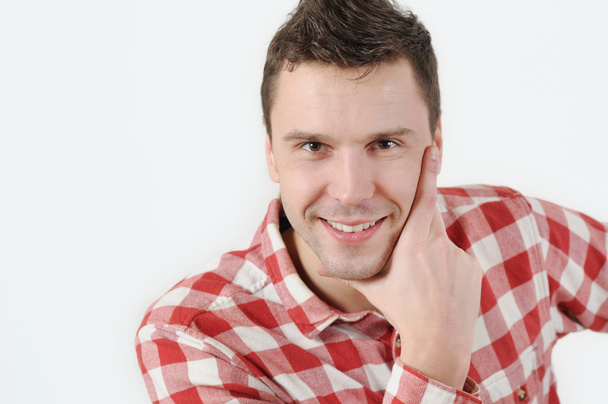 Smiling young man in hipster shirt holding hand on chin and standing against white background - Foto, immagini