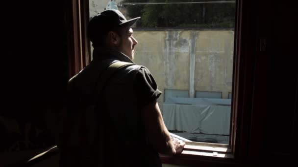Back side of man in cap watch in window. Inside building. Day. Bright sun - Filmati, video