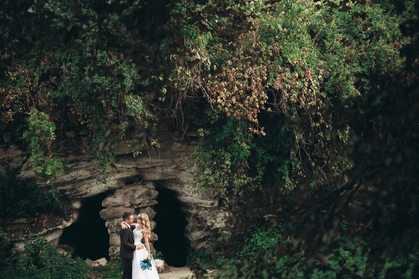 Luxus esküvői pár átölelve és megcsókolta a háttér gyönyörű növények, és a barlang ősi kastély közelében - Fotó, kép