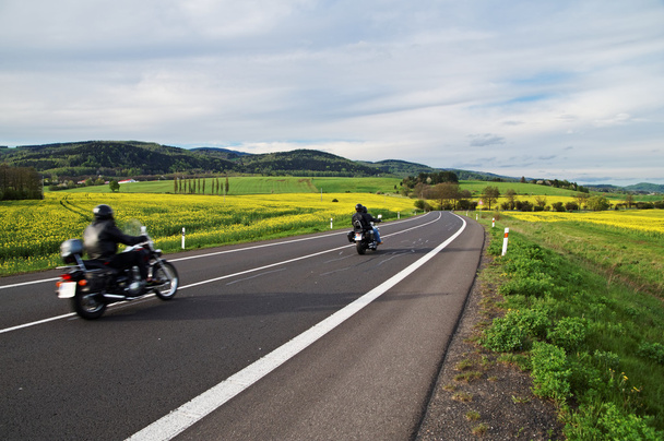 Motocicli che viaggiano lungo una strada asfaltata vuota tra campi di colza gialli in fiore
 - Foto, immagini