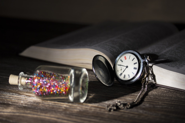 A small glass bottle with a bright colorful shiny beads and cork lid lies next to the old pocket watch and an open book illuminated by a light bulb. Photo in low key. Focus on watch - Foto, afbeelding