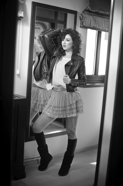 Young woman in black leather jacket and gray short tutu skirt looking into a large mirror. Beautiful curly dark hair girl posing in front of a wall mirror, indoors shot. Fashionable brunette model. - Foto, Imagem