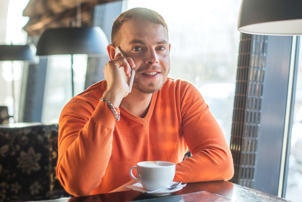 Handsome young man working, talking on the phone and smiling while enjoying coffee in cafe, look at camera - Фото, зображення