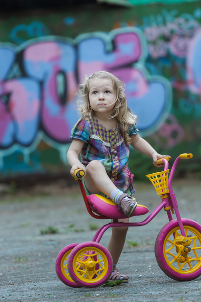 Kis kerékpáros lány visel kockás tunika lovaglás sárga és rózsaszín tricikli - Fotó, kép
