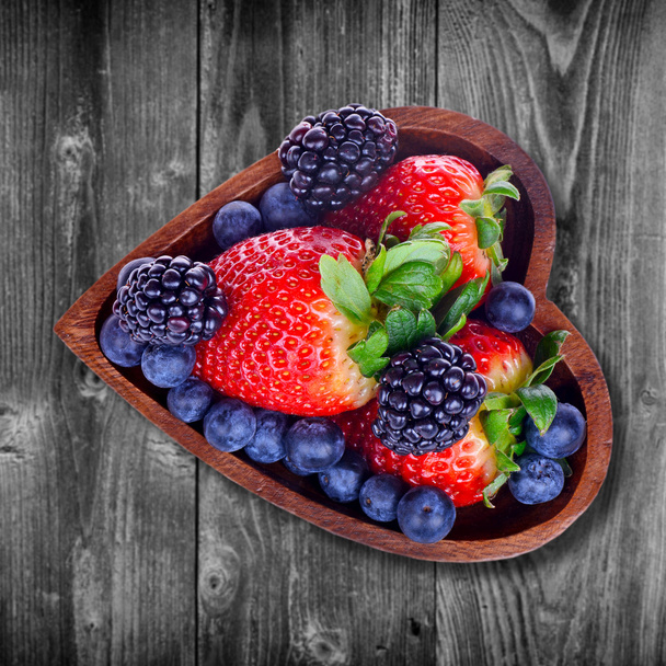 Various berries in a bowl in the form of heart on an old wooden surface - Zdjęcie, obraz
