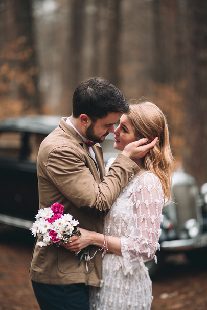 Romantic fairytale wedding couple kissing and embracing in pine forest near retro car. - Фото, изображение