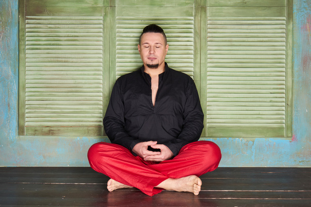 Portrait of a young man, black shirt and red slacks, hairstyle with shaved temples and slicked- back hair at the top of the head, different emotions, green wall, brown floor, beard, lotus posture - Foto, Imagen