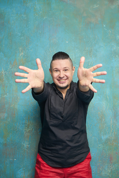 Portrait of a young man in red slacks and a black shirt, hairstyle with shaved temples and slicked- back hair at the top of the head, blue wall, beard, bright smiles and pulls the arms forward - Фото, изображение