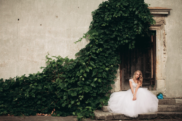 Beautiful young blond bride with bridal bouquet sitting on the stairs on the background gorgeous plants - 写真・画像