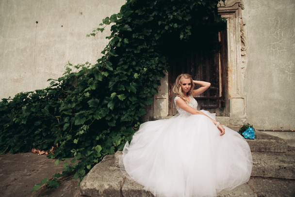 Beautiful young blond bride with bridal bouquet sitting on the stairs on the background gorgeous plants - Foto, Bild