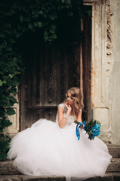 Szép fiatal szőke menyasszony, menyasszonyi csokor, ült a lépcsőn, a háttér gyönyörű növények - Fotó, kép