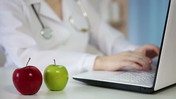 Female M.D. working in office, typing on laptop. Apples on table, healthy eating - Video