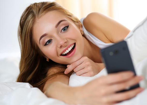 Jonge mooie blonde vrouw liggend in bed holding cellphone - Foto, afbeelding