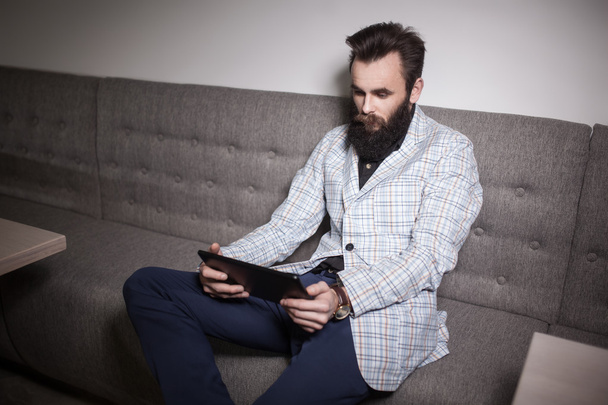 bearded man in suit and with tablet sitting in cafe and browsing - Фото, изображение