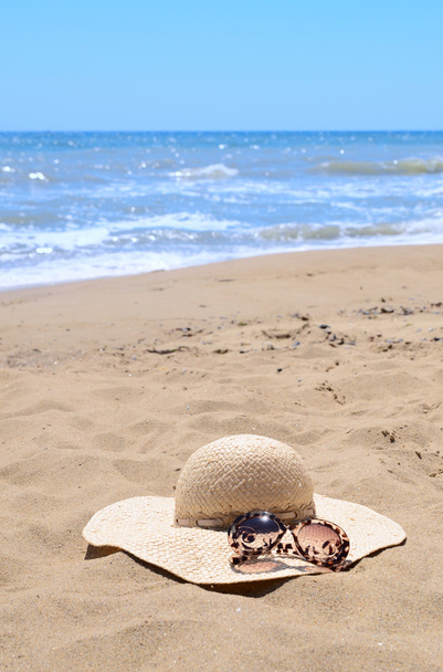 Zonnebrillen en stro hoed liggend op het zand op een strand tegen de zee - Foto, afbeelding