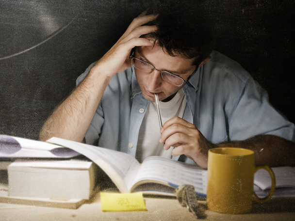 Young student at home desk reading biting pen studying at night with pile of books and coffee - Photo, Image
