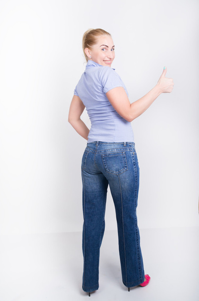 hermosa joven rubia de piernas largas en un jeans y una camisa azul en botas, posando en altura completa, muestra el pulgar
 - Foto, Imagen