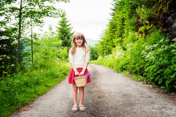 Чарівна дівчинка 6-7 років ходить в лісі, тримає невеликий кошик з квітами, носить бежевий пуловер і червону спідницю
 - Фото, зображення