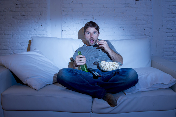 man sitting at home sofa in living room watching movie or sport in tv eating popcorn and drinking beer - Fotó, kép