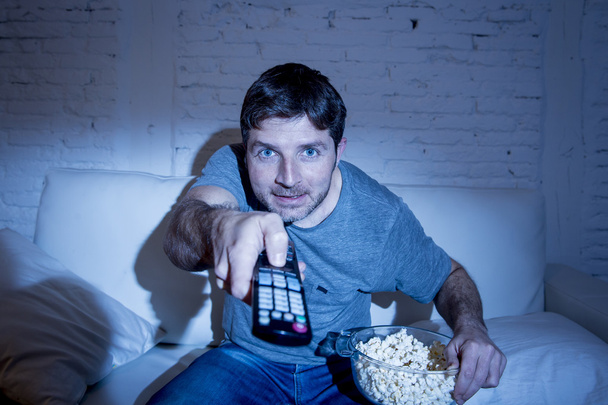 man at home lying on couch at living room watching tv eating popcorn bowl using remote control - Photo, Image