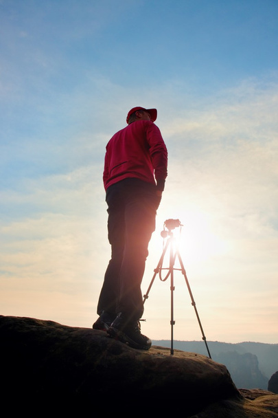 Fotograf pobyt z statywu na klif i myślenia. Senny fogy krajobraz, pomarańczowy mglisty wschód słońca w pięknej dolinie - Zdjęcie, obraz