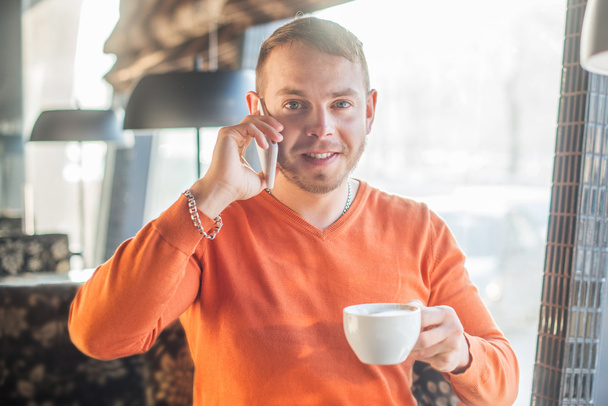 Handsome young man working, talking on the phone and smiling while enjoying coffee in cafe, look at camera - Foto, Imagen