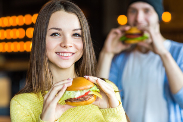 Man and woman eating burger. Young girl and young man are holding burgers on hands - Фото, зображення