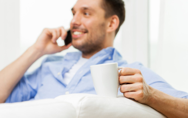 smiling man with cup calling on smartphone at home - Фото, изображение