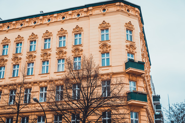 Низкоугловой архитектурный экстерьер жилого здания с низким подъемом с жёлтым фасадом и классическими скульптурными деталями и балконами в Малый уголок в пасмурный день с голыми деревьями
 - Фото, изображение