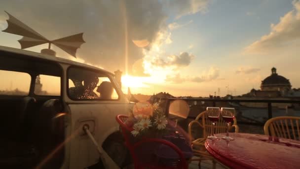 Young loving couple kissing in the vintage car on the rooftop cafe with glasses of red wine and flowers on the table. View of ancient city building while raining. Romantic sunset on the background - Footage, Video