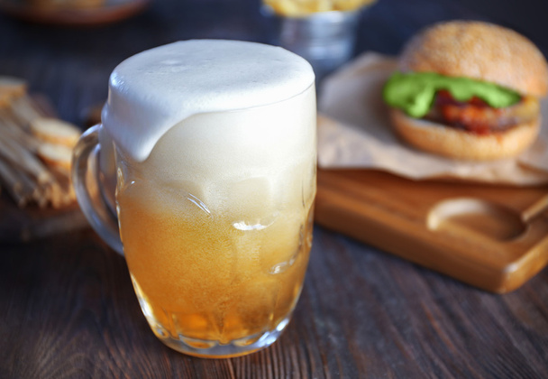 Glass mug of light beer with hamburger on dark wooden table, close up - Фото, изображение