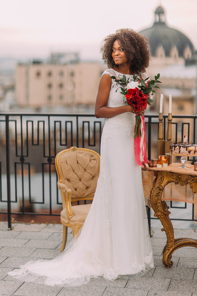 Charming black bride with wedding bouquet in hands standing on the terrace - Fotó, kép