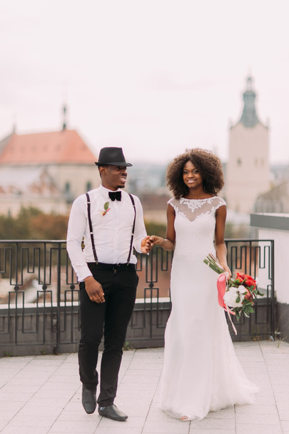 Lovely african wedding couple holding hands on the roof with view on Lviv city architecture - Foto, Imagen