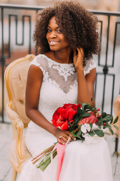 Happy young black bride smiling with bouquet of red flowers and sitting on vintage terracotta chair - Foto, Bild