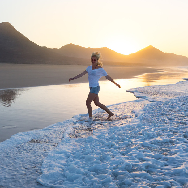 Κυρία που απολαμβάνουν τρέχει από κύματα σε αμμώδη παραλία στο ηλιοβασίλεμα. - Φωτογραφία, εικόνα