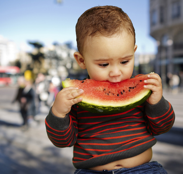 Portrait of a handsome kid biting a watermelon against at a crow - Photo, Image