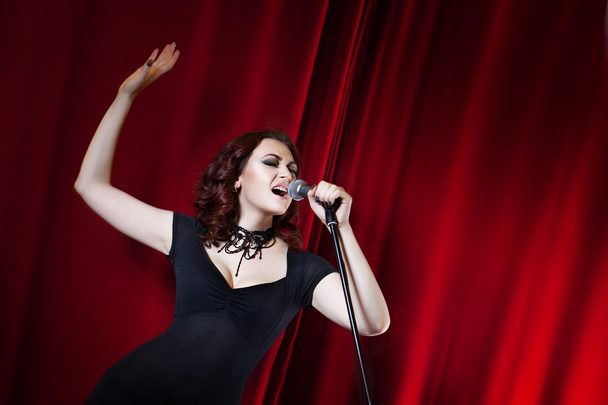 Beautiful Singing Girl on stage. Beauty Woman with Microphone. Red curtain  background - Fotó, kép