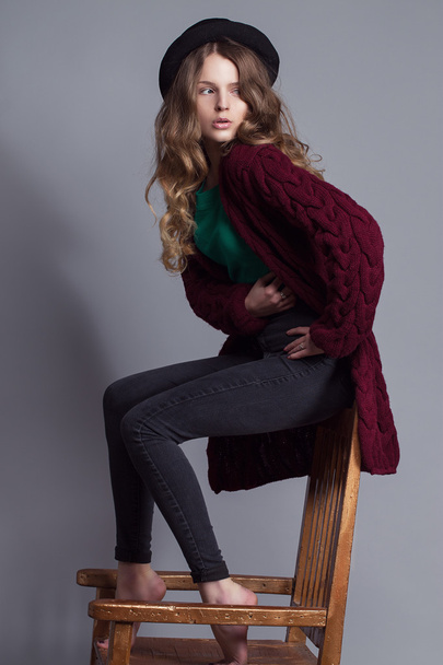 Modeporträt einer jungen schönen Frau in Freizeitkleidung (Strickjacke, Jeans und grünem T-Shirt). Ohne Stiefel, mit Hut und natürlichem Make-up sitzt sie auf einem Stuhl. Kopierraum. - Foto, Bild