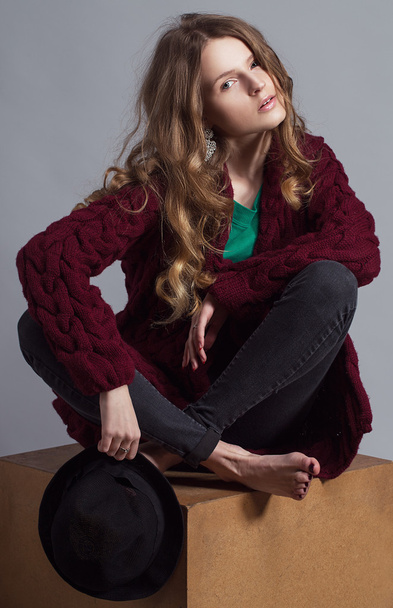 Modeporträt einer jungen schönen Frau in Freizeitkleidung (Strickjacke, Jeans und grünem T-Shirt). Ohne Stiefel, mit Hut und natürlichem Make-up sitzt sie auf einem Holzwürfel. Kopierraum. - Foto, Bild