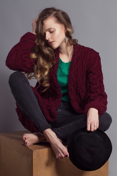 Fashion portrait of young beautiful woman model in casual wear (knitted cardigan, jeans and a green t-shir). without boots, in hat and natural make up on her face sitting on a wooden cube. Copy space. - Photo, Image